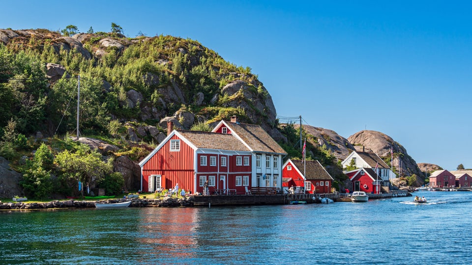 Red and white houses in Norway, right on the water.  Behind there are bushes and rocks.