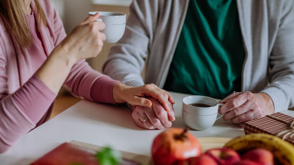 Woman on the left (only upper body and arms in picture) and older man (right, also only upper body and arms) are drinking coffee