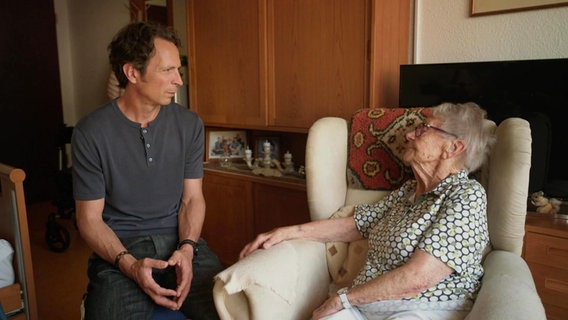 Moderator Jo Hiller talks to an older woman who is sitting in a wing chair in a nursing home apartment.  © Screenshot 