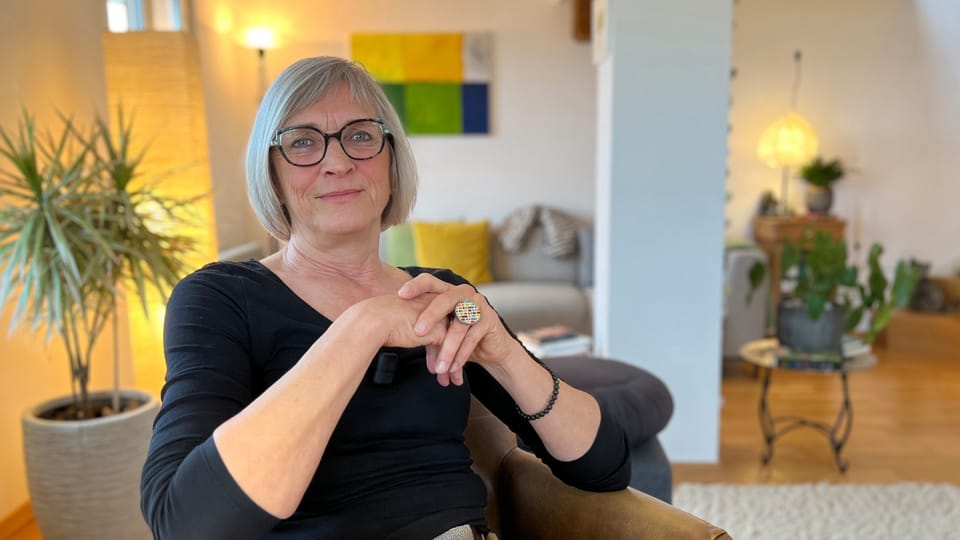 Woman sitting at a table, gray, medium-length hair, black, large glasses.  In the background the living room with sofa and light