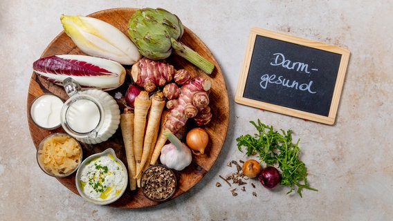 Arranged on a round plate are root vegetables, artichoke, chicory, onion, garlic, kefir, herb quark, kimchi and spices.  Next to it is a board: "Gut healthy" © NDR Photo: Claudia Timmann