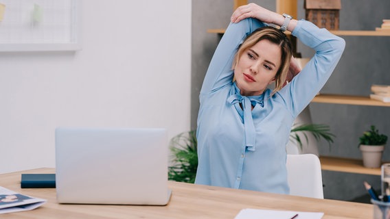 A young woman sits at her desk and does stretching exercises. © PantherMedia Photo: IgorVetushko