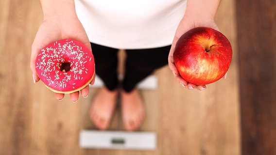 Woman stands on a scale and holds a doughnut and an apple in her hands © Fotolia.com Photo: Maksymiv Iurii