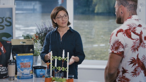 Doc Schäfer shows her muscular patient a model of a chemical factory, next to which are various protein powders. © NDR Photo: Oliver Zydek/Moritz Schwarz