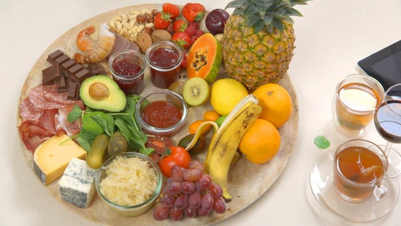 A large plate with various nuts, strawberries, banana, tomatoes, gherkins, ketchup, blue cheese, papaya, pineapple, sauerkraut, citrus fruits, kiwi, chocolate, jams, crustaceans and plums, next to green tea and red wine. © NDR 