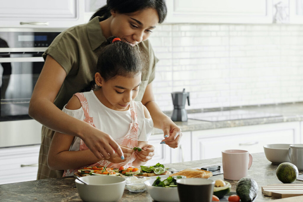 A woman and a child are putting various types of bread and vegetables on the kitchen counter.