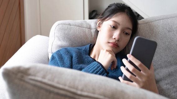 A young girl sitting on the sofa looks worriedly at her smartphone © wichayada (YAYMicro) / Panthermedia Photo: wichayada (YAYMicro) / Panthermedia